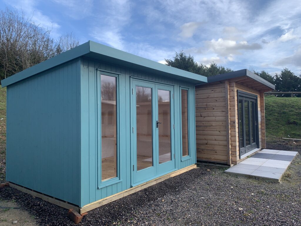 Two modern pent-roof garden rooms, one painted teal and one natural timber with large glass doors, on display near Harlow.